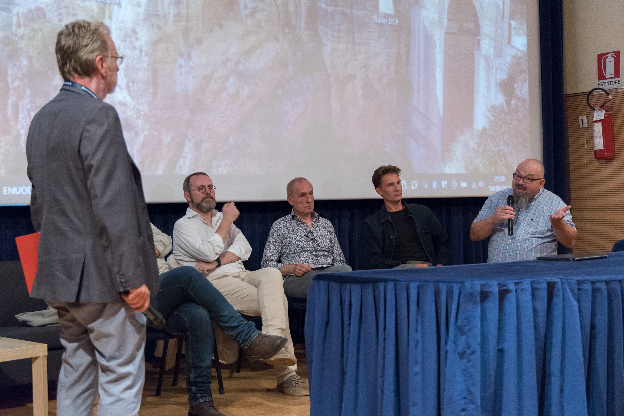 Tavolo dei relatori con moderatore in piedi e domanda dal pubblico